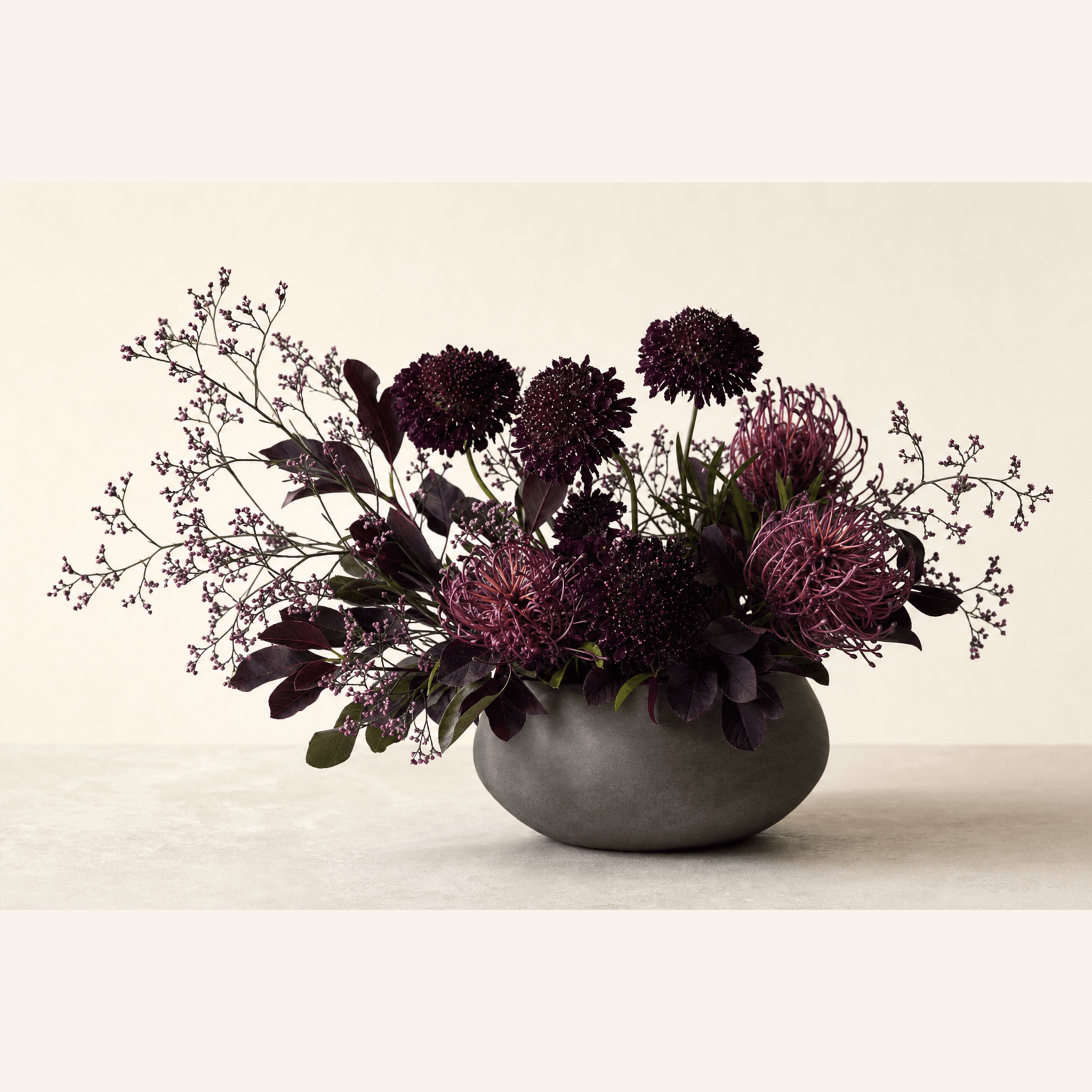 Deep purple scabiosa and pink pincushion protea with limonium in a dark stone bowl