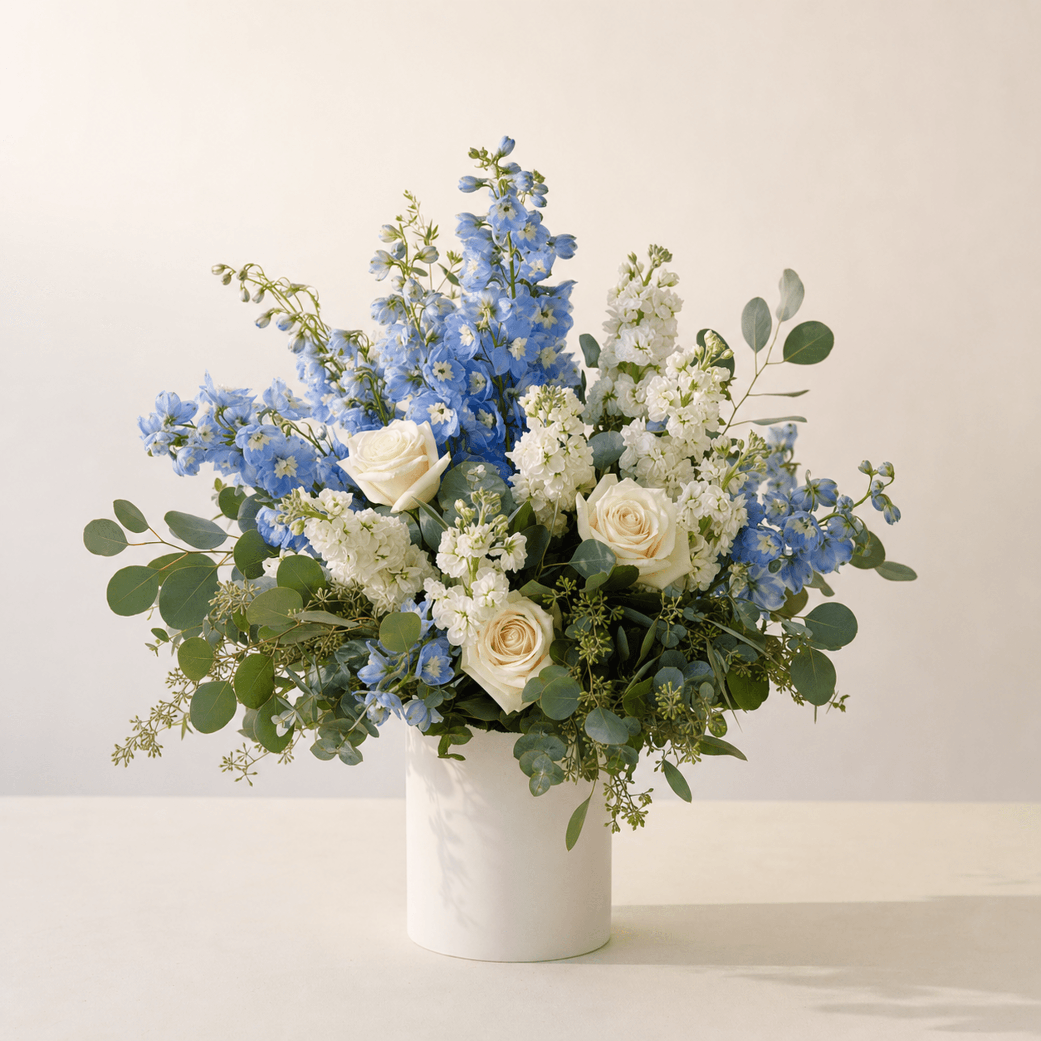 Blue delphinium, white stock, cream roses and eucalyptus in a white marble cylinder vase