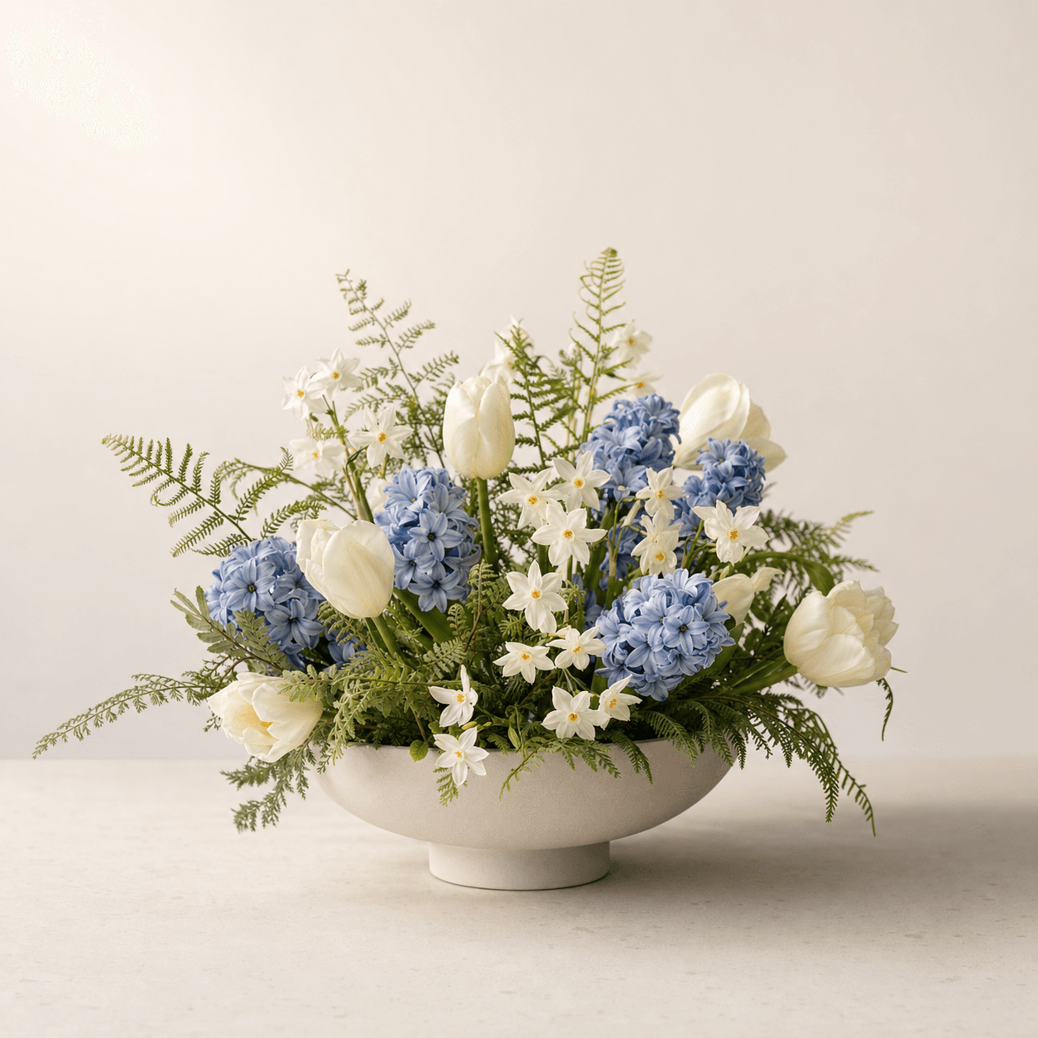 Blue hyacinths, white tulips and white narcissi arranged with fern fronds in a cream ceramic footed bowl