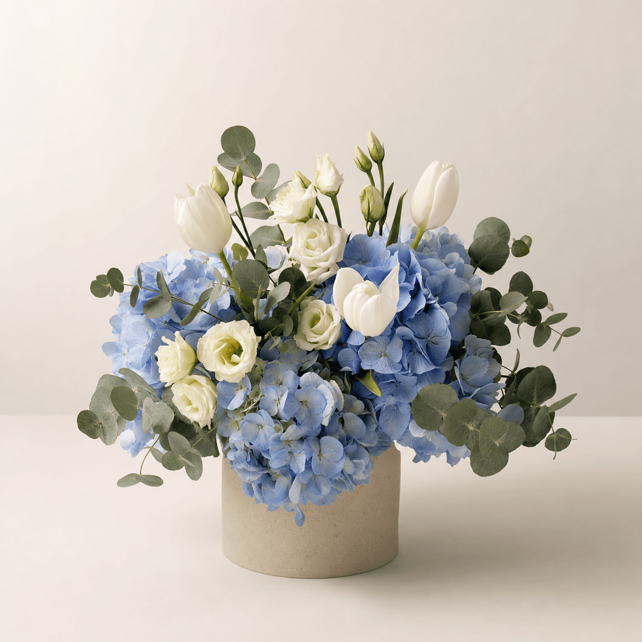 Blue hydrangeas, white tulips, cream lisianthus and silver eucalyptus in a cylindrical stone vessel
