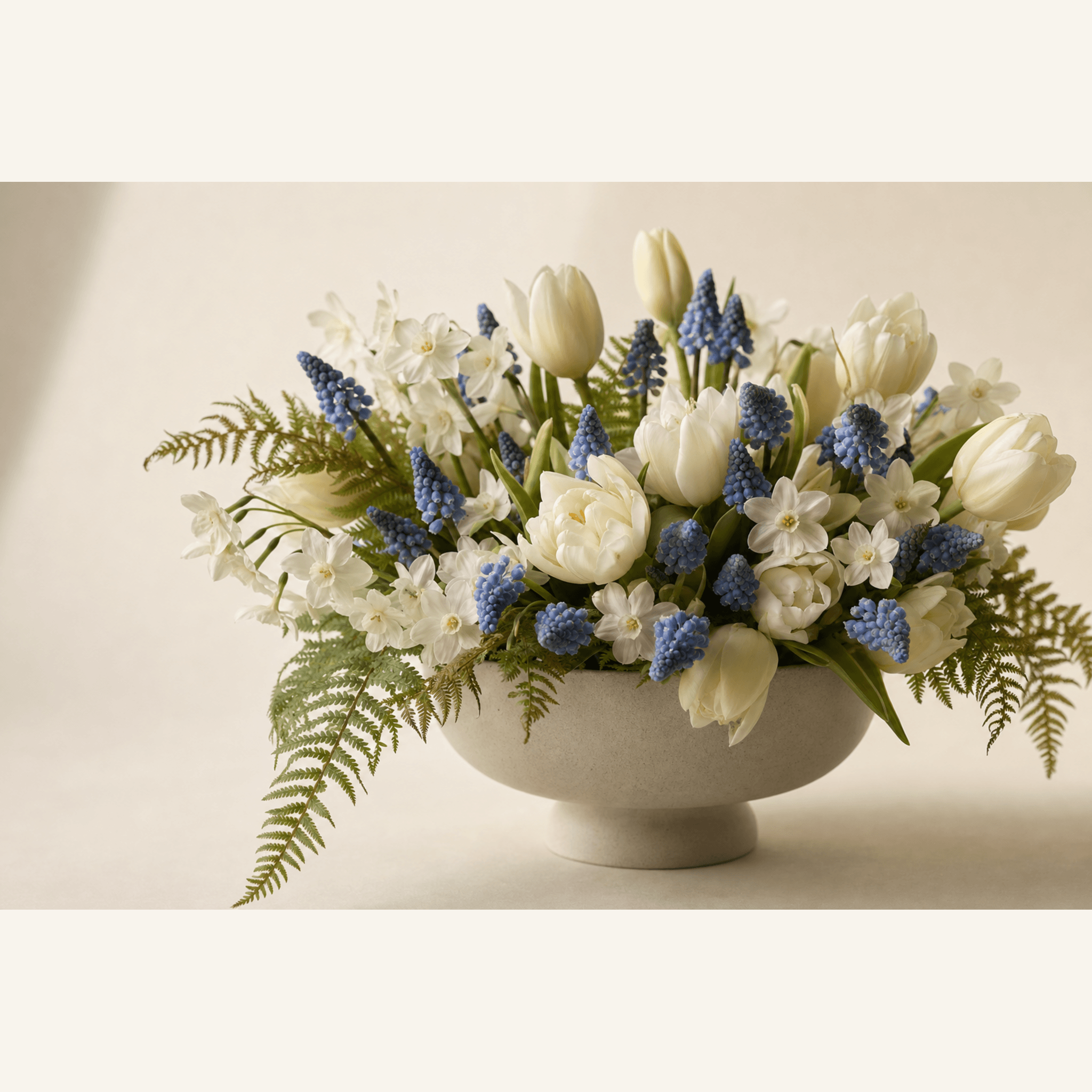 White tulips, blue muscari, white narcissus and fern fronds arranged in a cream ceramic pedestal bowl