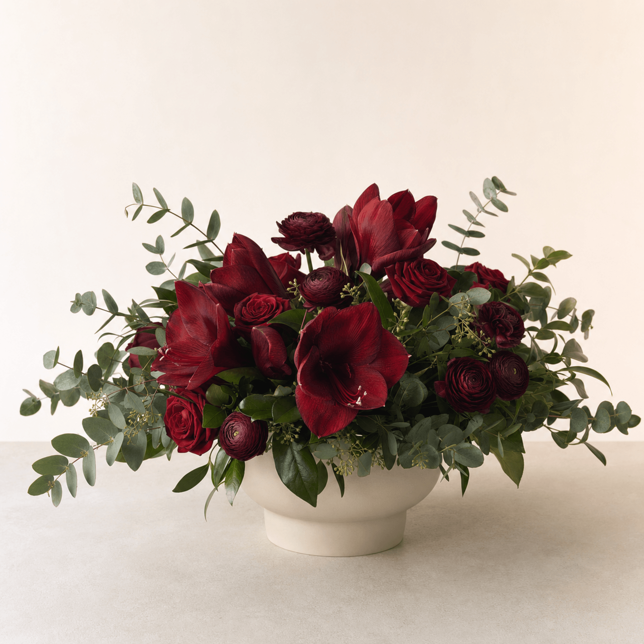 Deep red amaryllis, roses and ranunculus with eucalyptus in a cream ceramic bowl