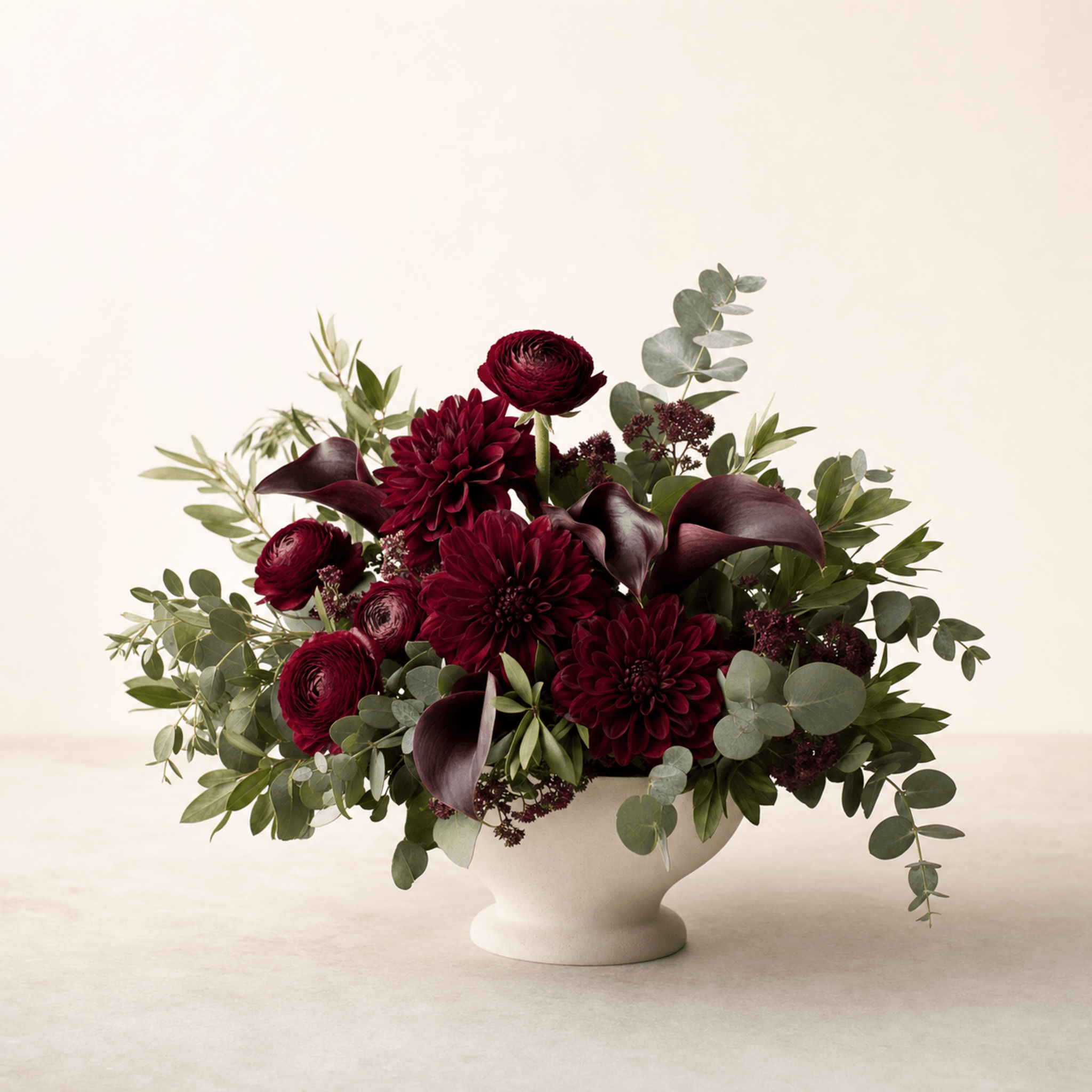 Deep crimson dahlias, burgundy ranunculus and dark calla lilies in a white pedestal bowl