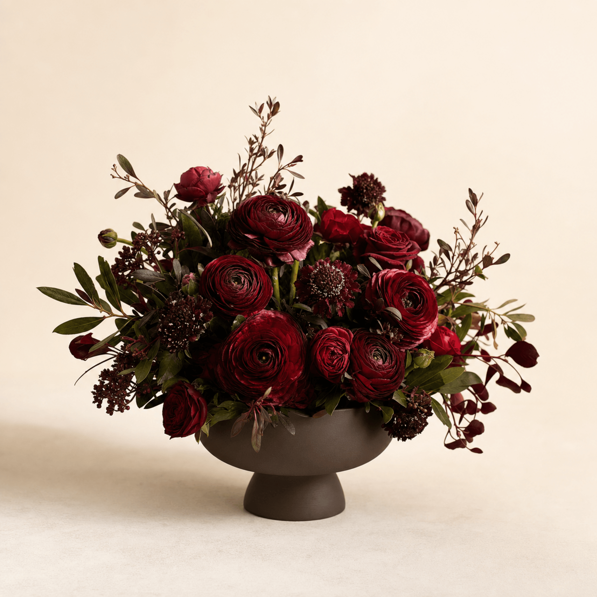 Deep crimson ranunculus, dark red roses and burgundy scabiosa arranged in a dark matte footed bowl