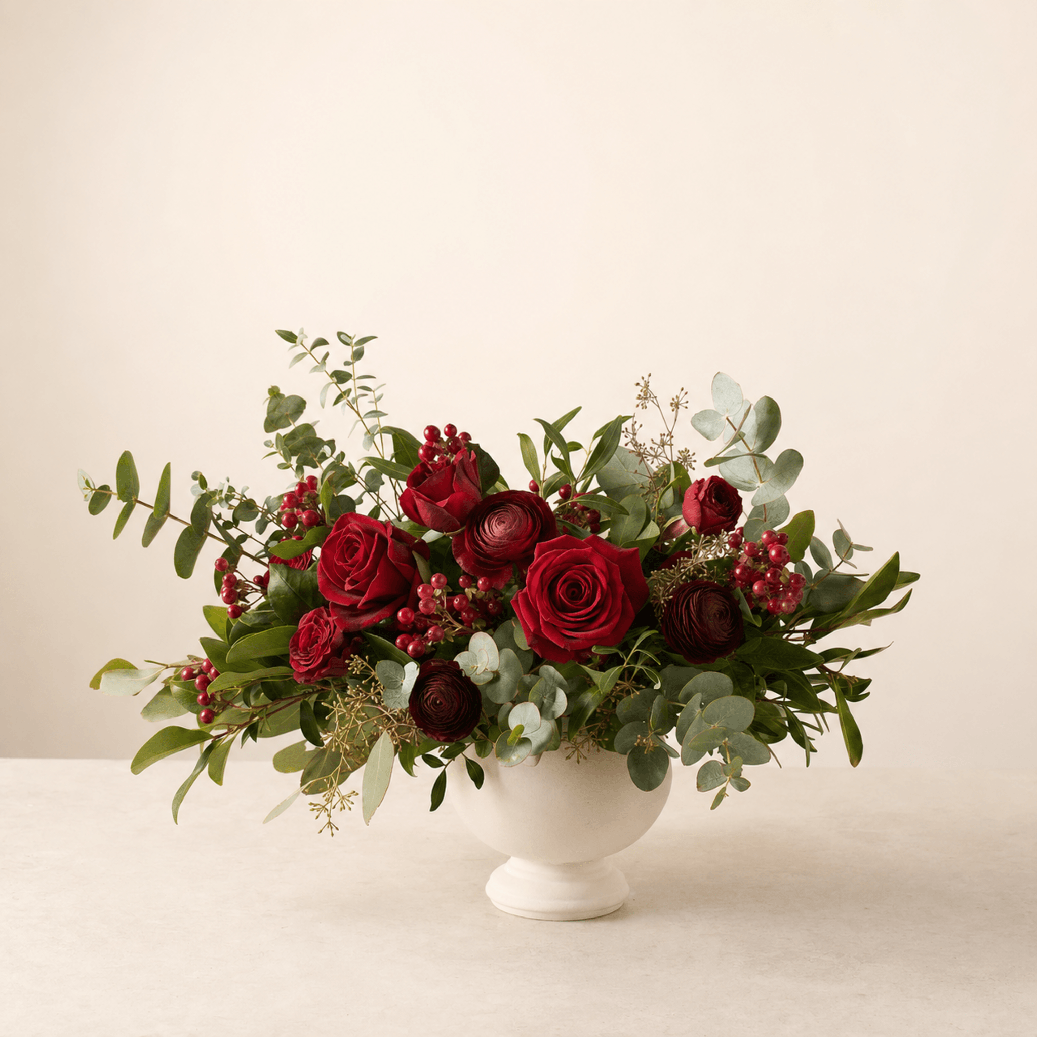 Deep red roses and burgundy ranunculus with eucalyptus and red berries in a white footed bowl