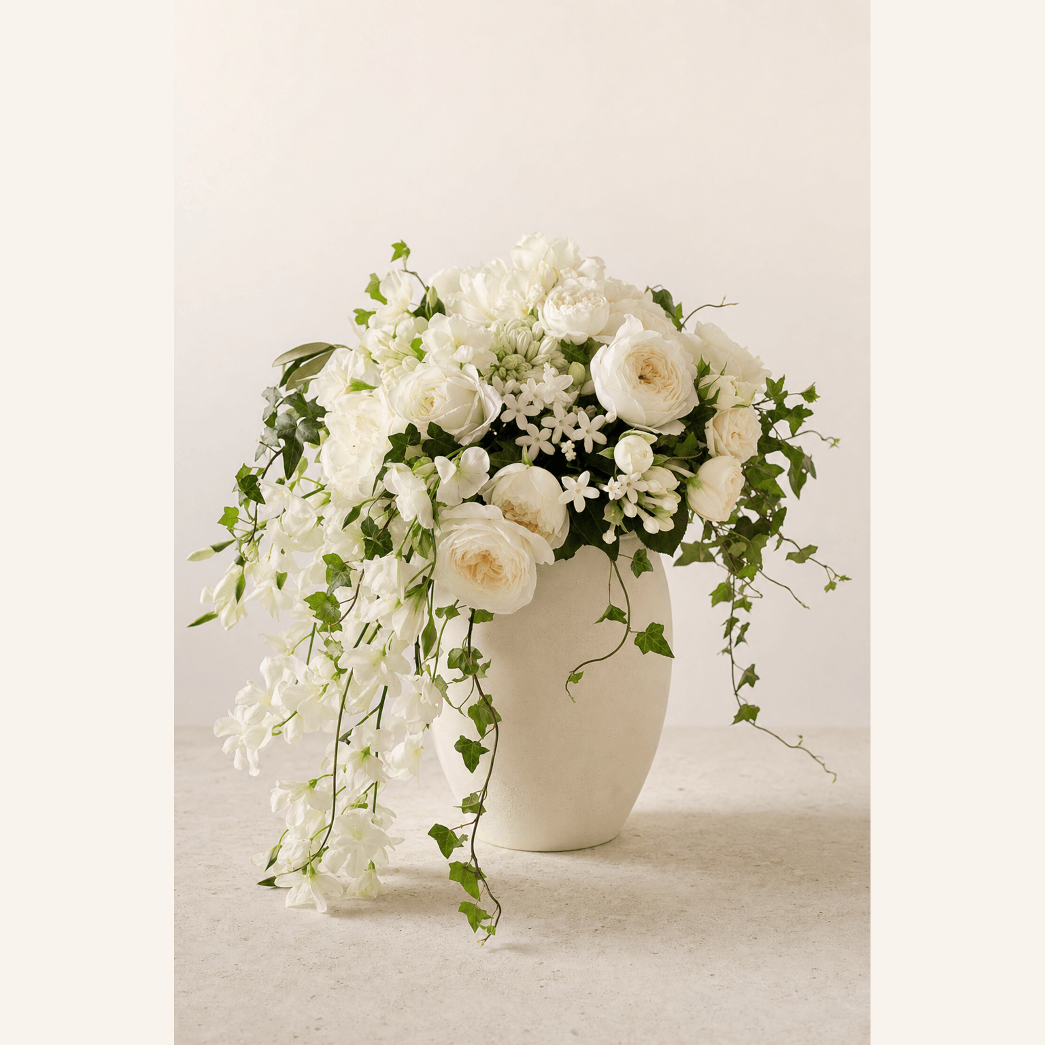 White garden roses, peonies, freesia and ivy trailing from a matte cream ceramic vase