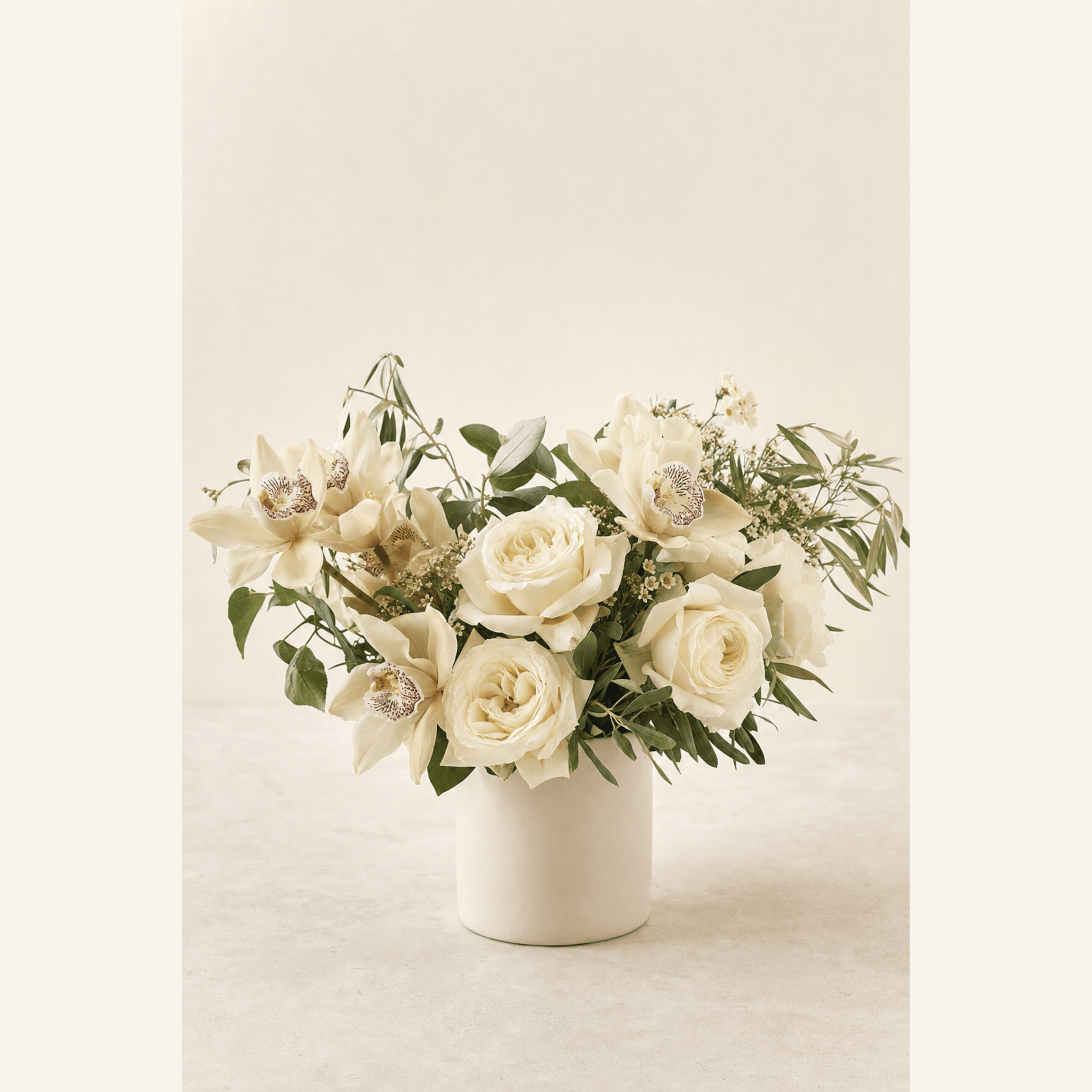 Cream roses and white cymbidium orchids with soft greenery in a white ceramic cylinder vase