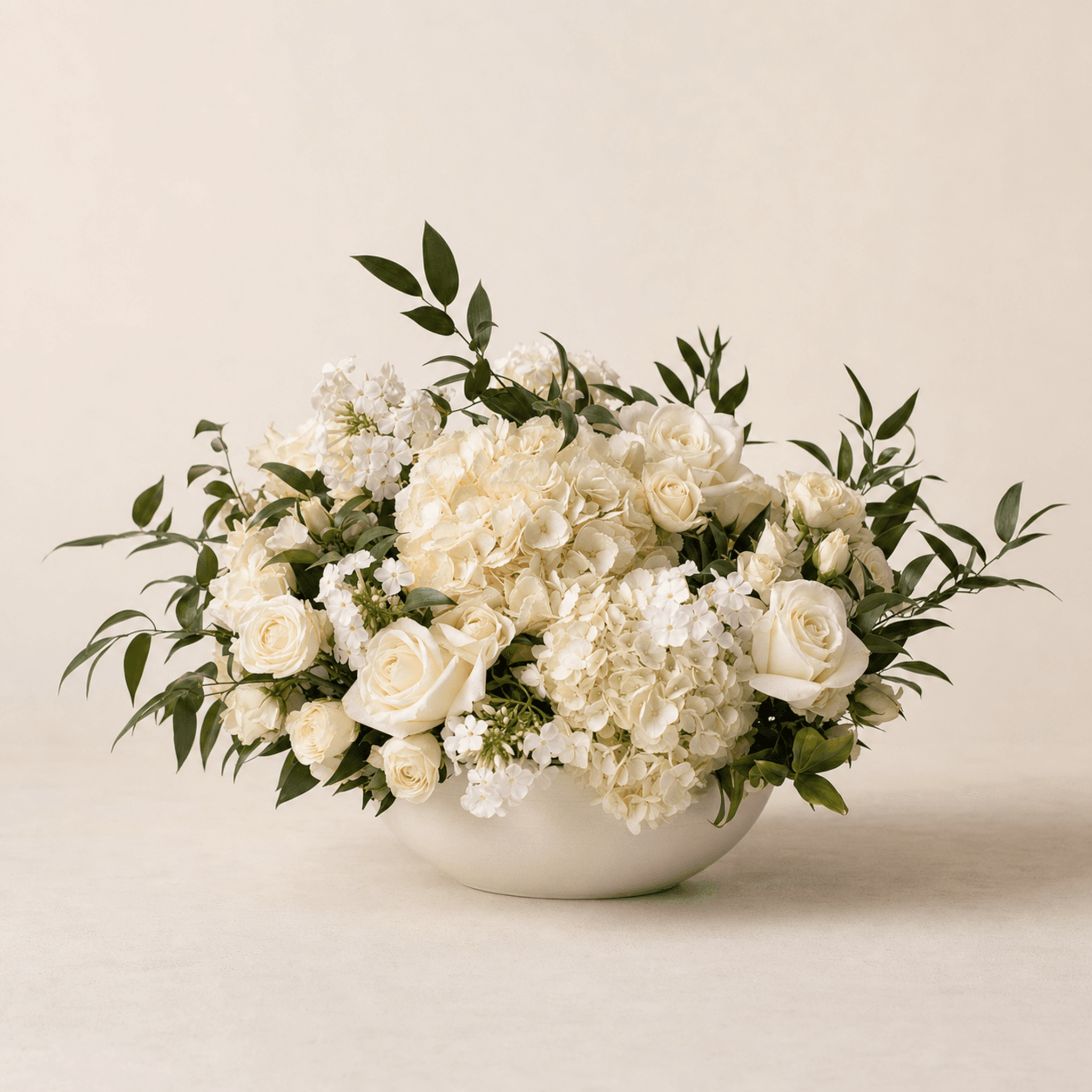 Cream roses, white hydrangeas and stock arranged in a wide white ceramic bowl with trailing greenery