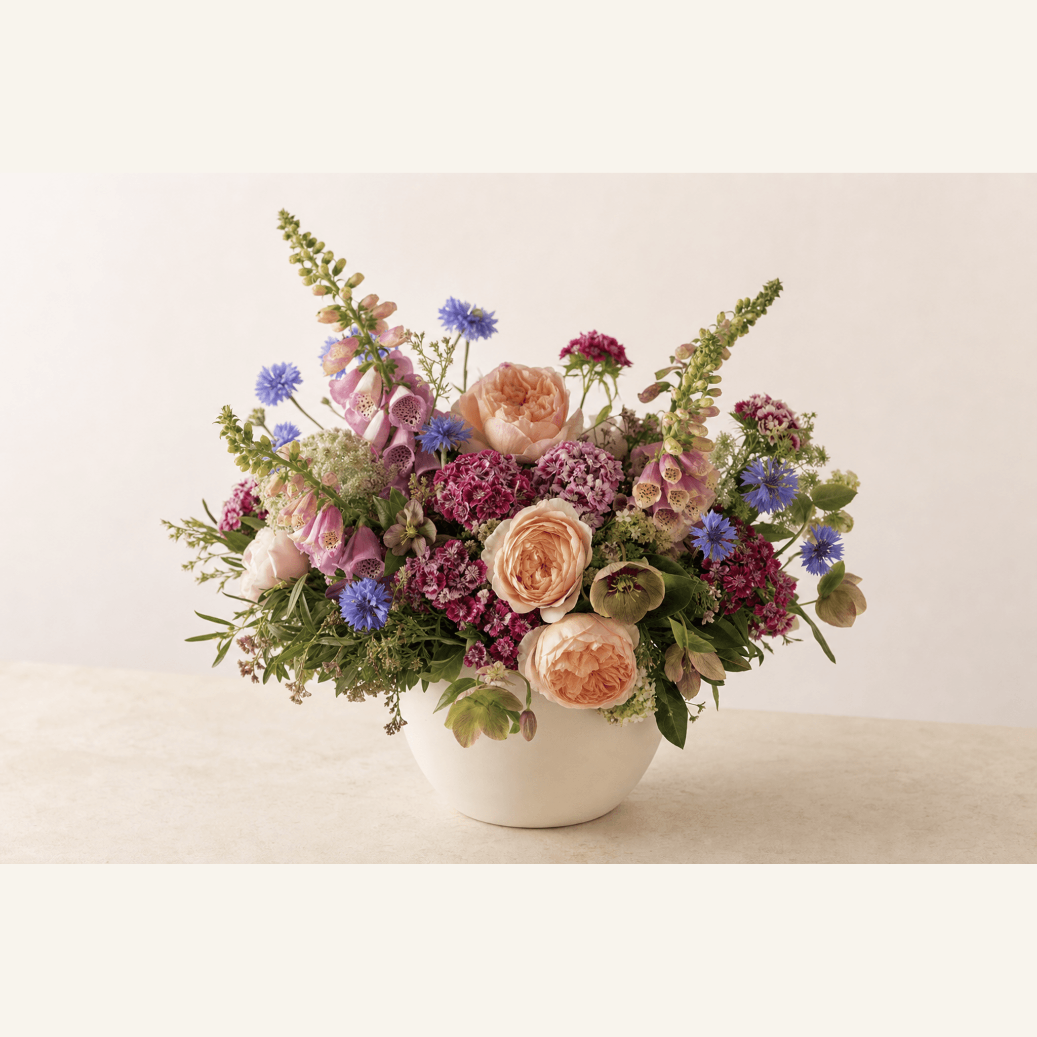 Peach garden roses, pink foxgloves, blue cornflowers, and magenta sweet Williams in a white ceramic bowl