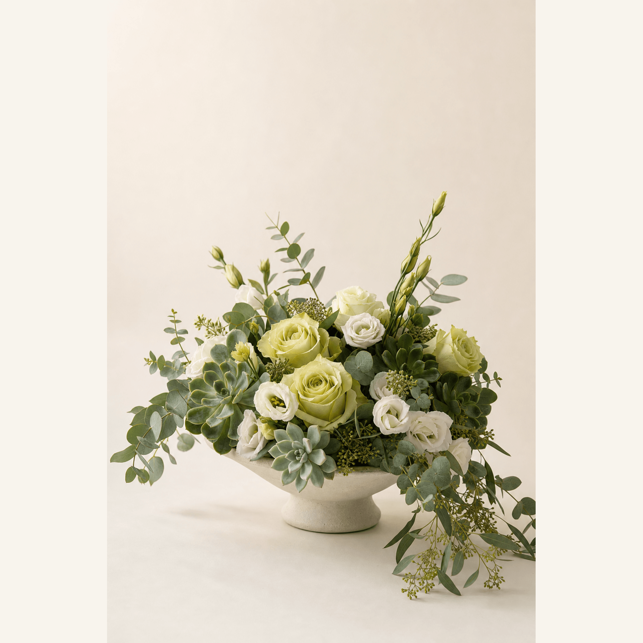 Pale green and white roses, lisianthus, eucalyptus and succulents arranged in a cream ceramic footed bowl