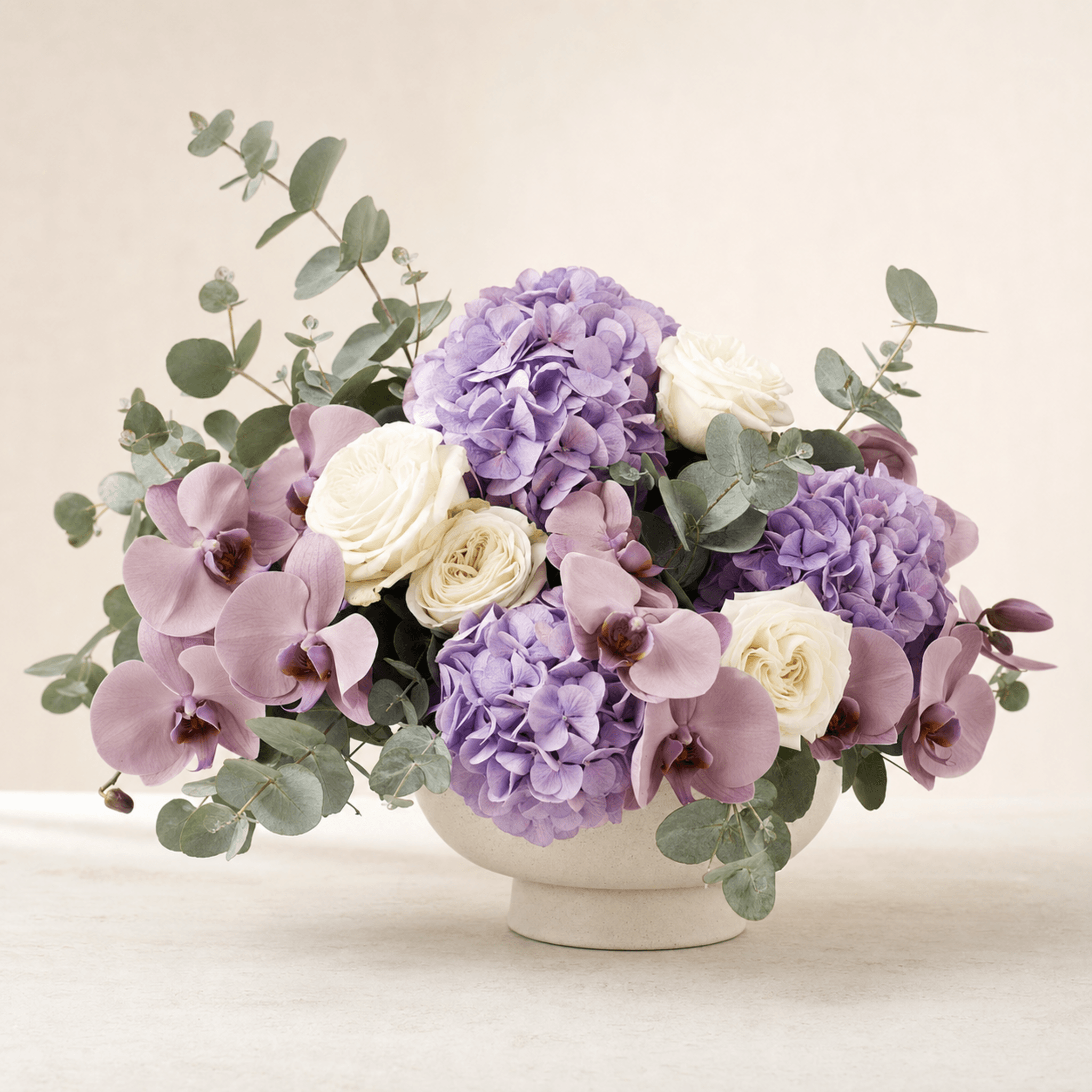 Purple hydrangeas, mauve orchids and cream roses arranged in a wide cream ceramic bowl with eucalyptus