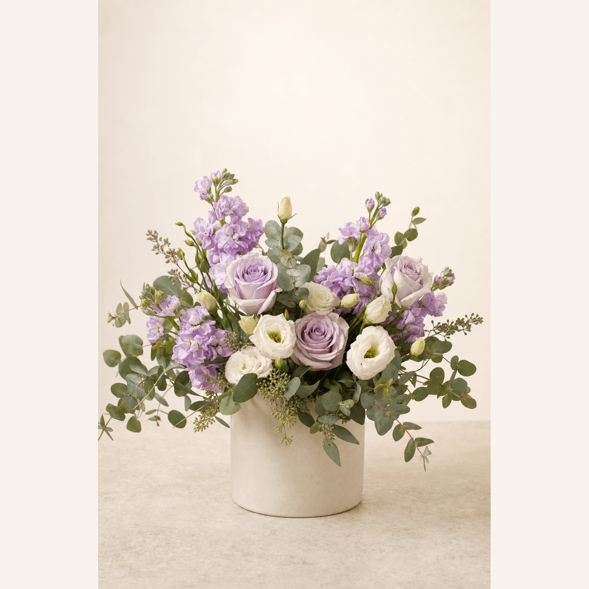 Lavender roses, white lisianthus and purple stock in a cream ceramic pot with eucalyptus