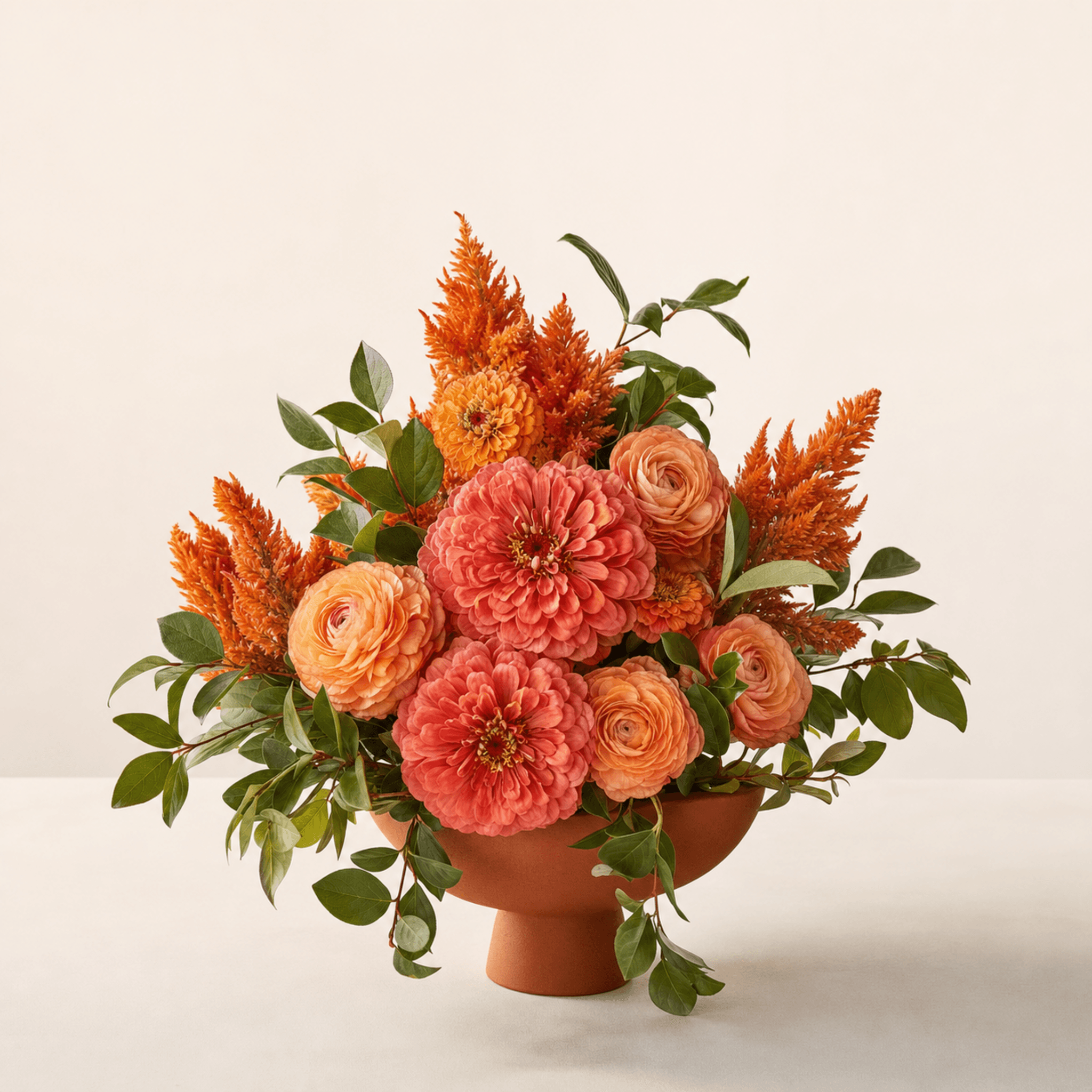Coral zinnias, peach ranunculus and orange celosia arranged in a terracotta footed bowl with trailing greenery