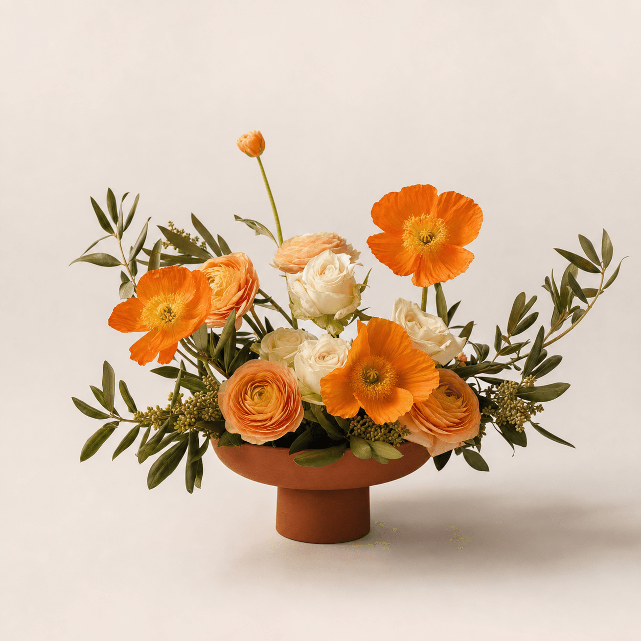 Orange poppies, peach ranunculus and white roses arranged in a terracotta pedestal bowl with olive foliage