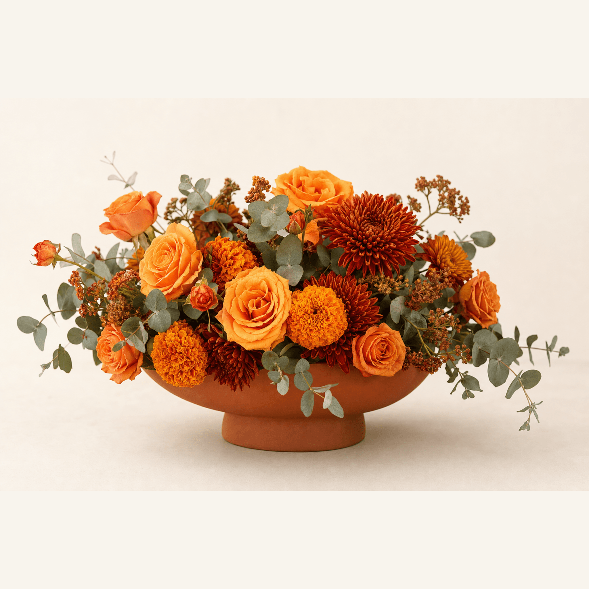 Orange roses, marigolds, rust chrysanthemums and eucalyptus in a terracotta footed bowl