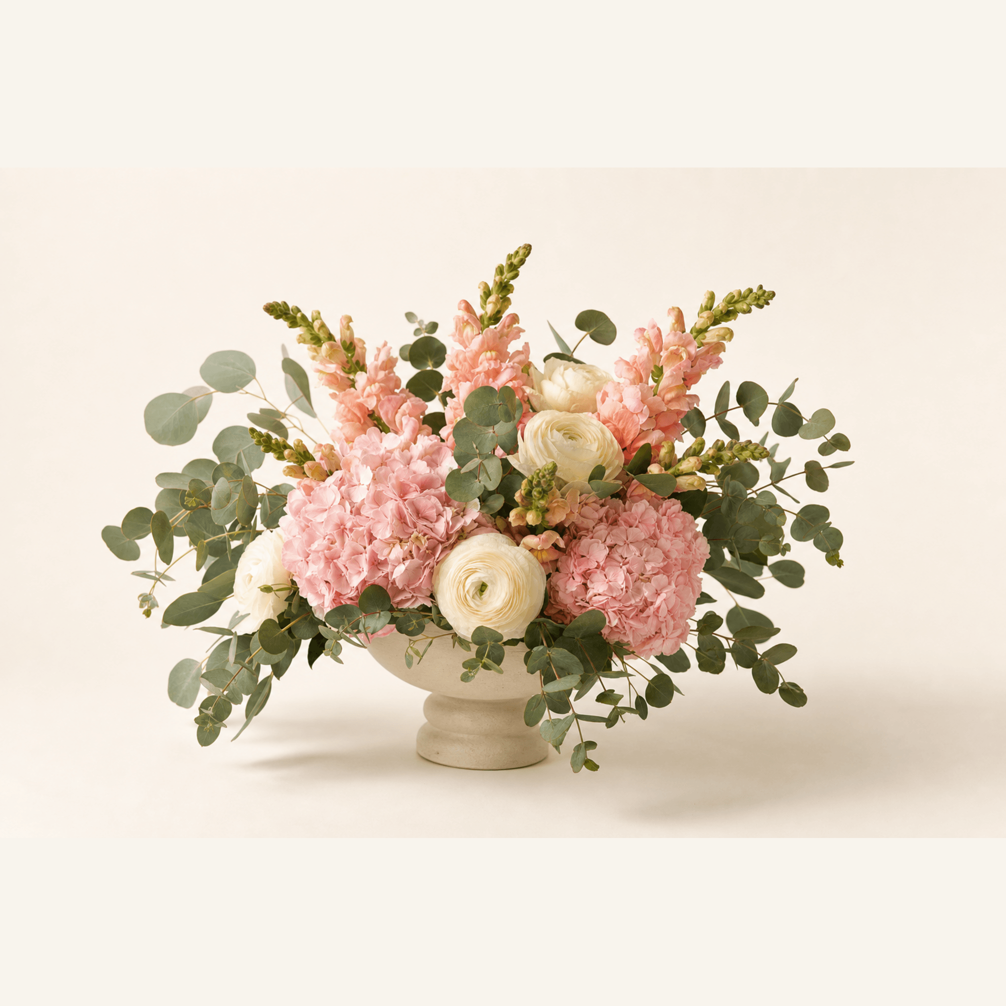 Pink hydrangeas, white ranunculus and peach snapdragons arranged in a cream pedestal bowl with eucalyptus