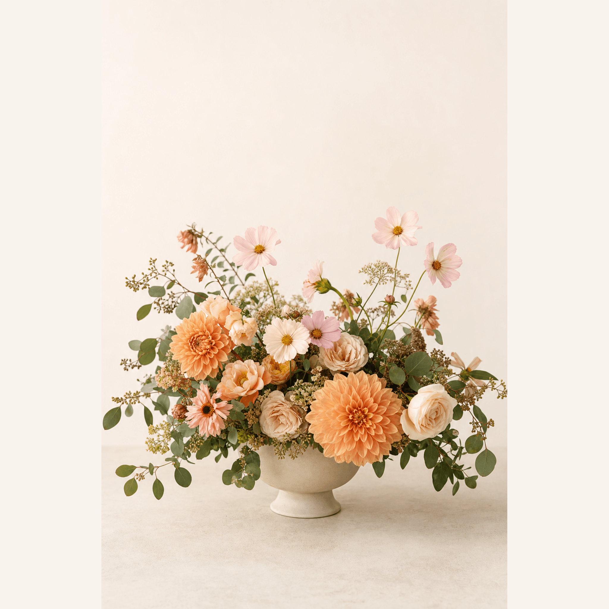 Peach dahlias, cream roses, pink cosmos and eucalyptus arranged in a cream ceramic footed bowl