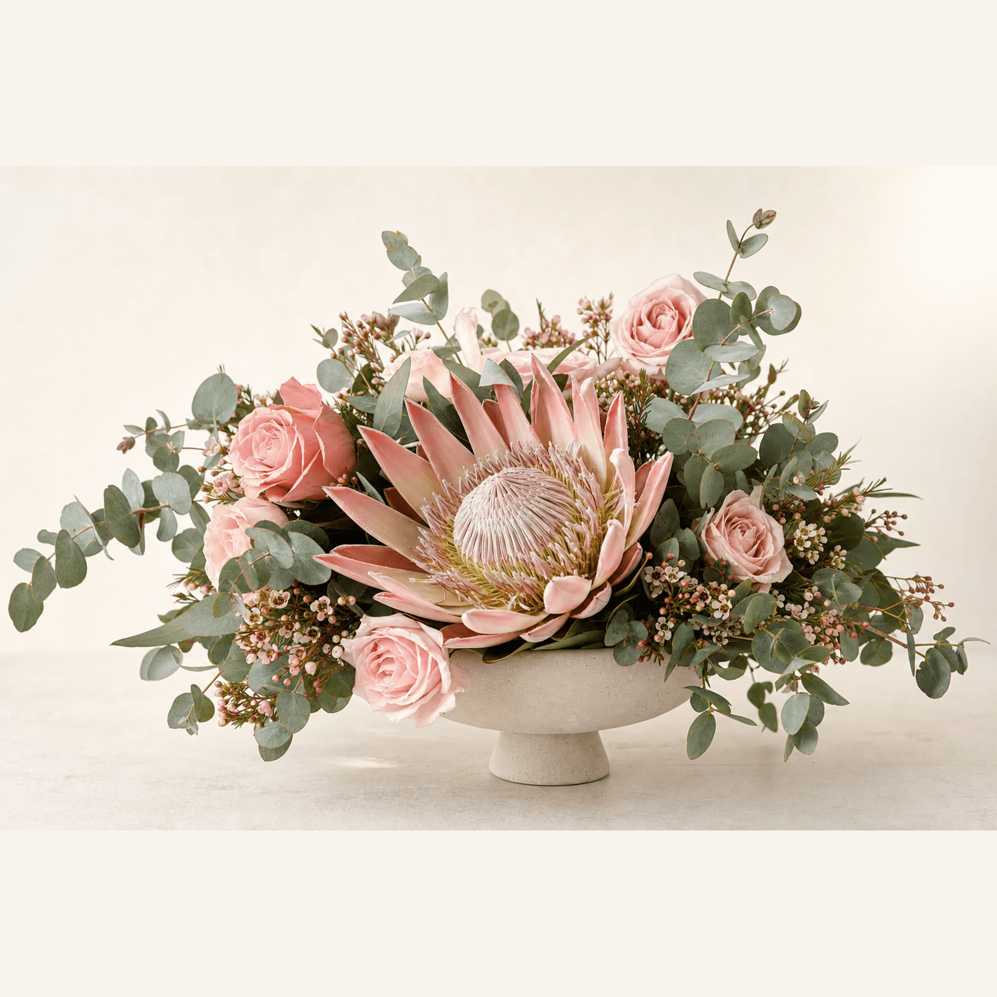 Pink king protea, blush roses, wax flower and eucalyptus in a white footed ceramic bowl