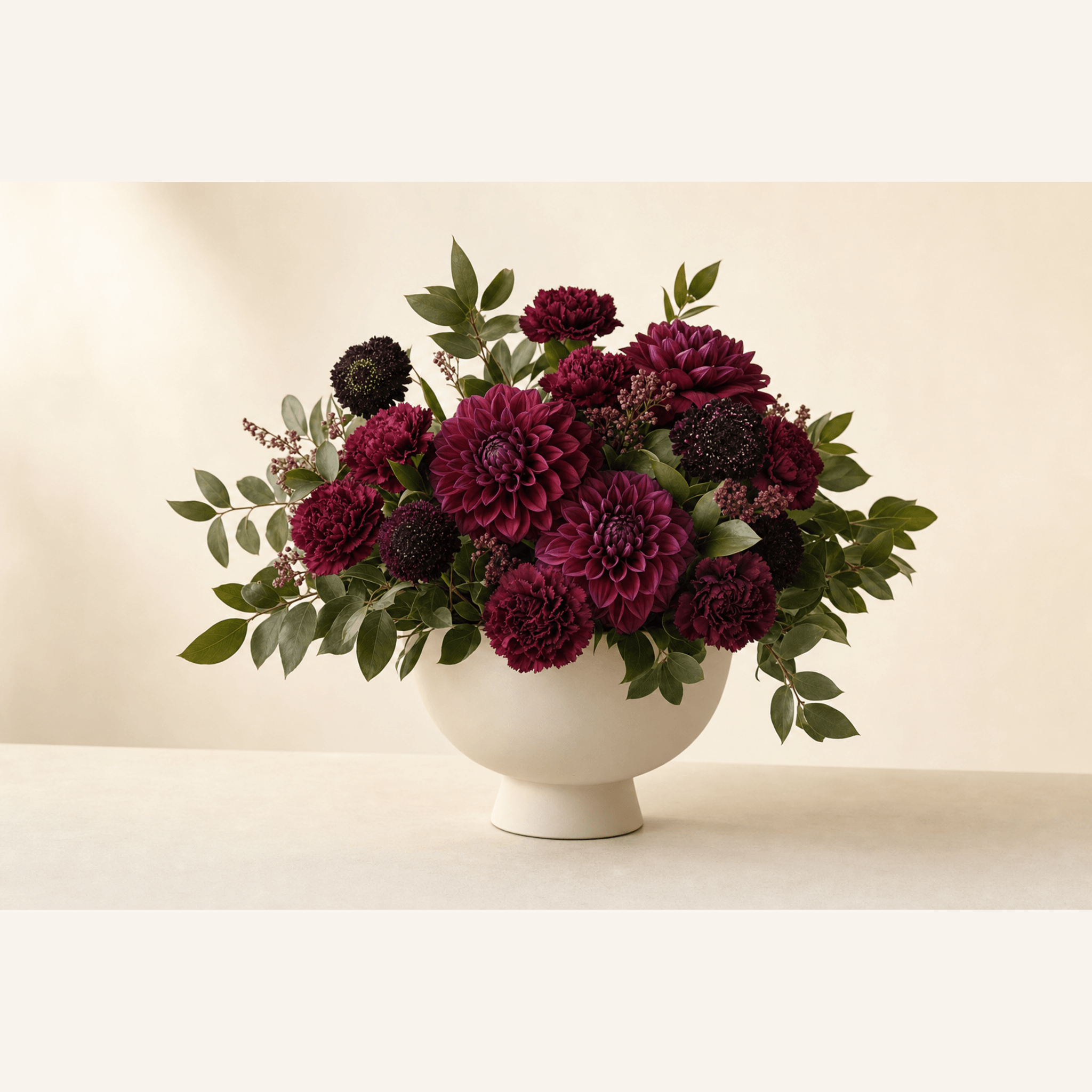 Deep burgundy and purple dahlias, carnations and scabiosa in a cream ceramic pedestal bowl
