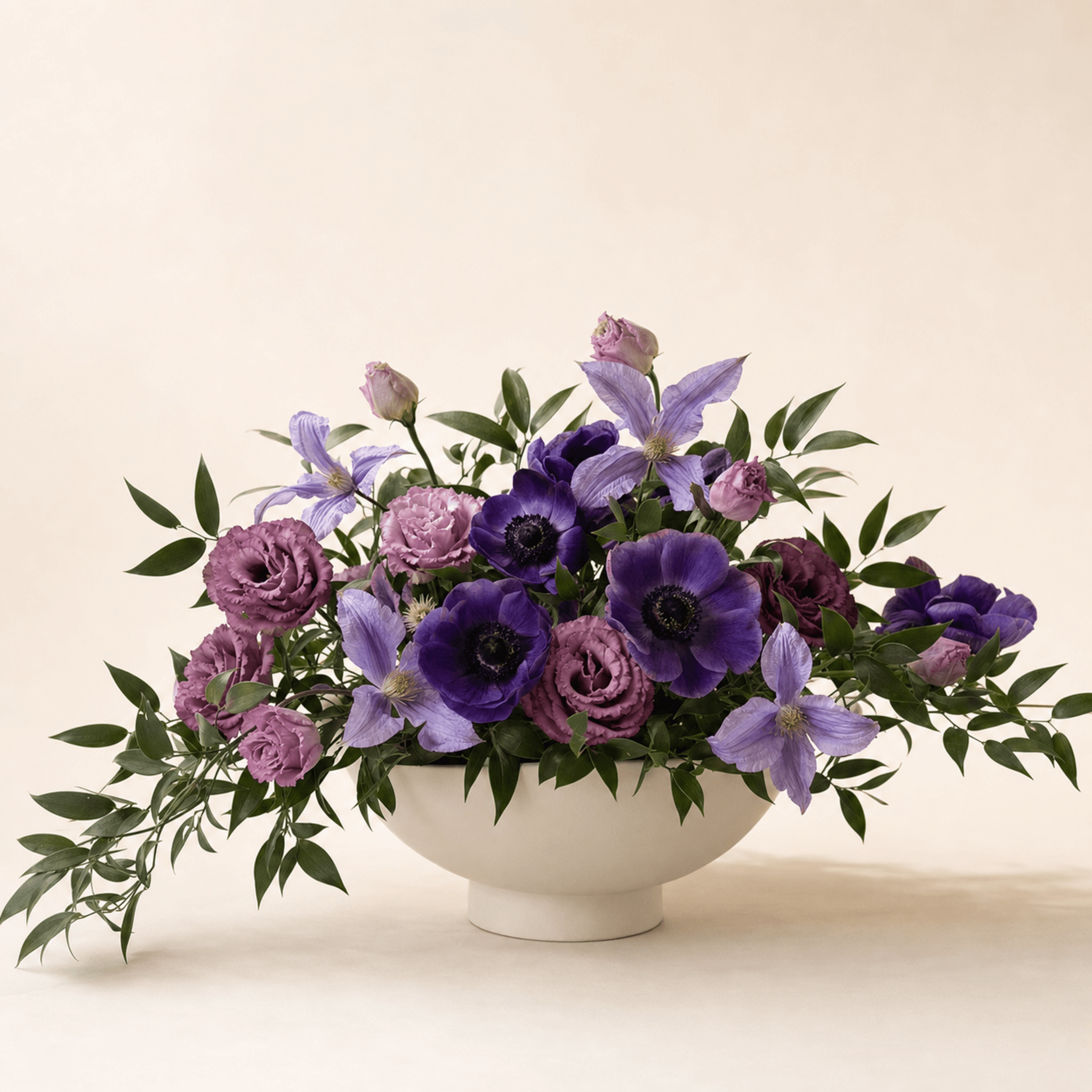 Deep purple anemones, lilac clematis and dusty-pink lisianthus arranged in a wide white ceramic bowl
