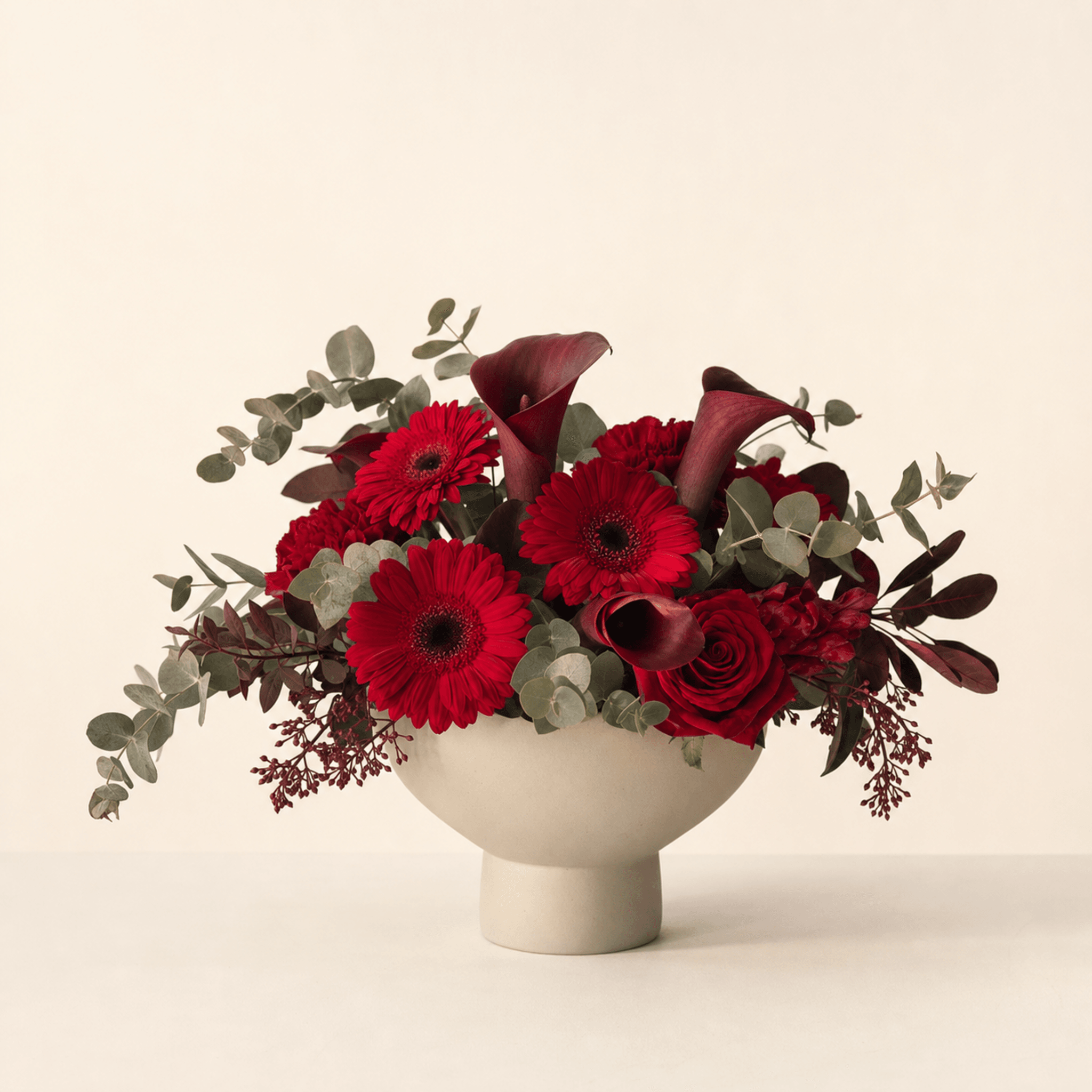 Red gerberas, deep burgundy calla lilies and red roses with eucalyptus in a cream pedestal bowl