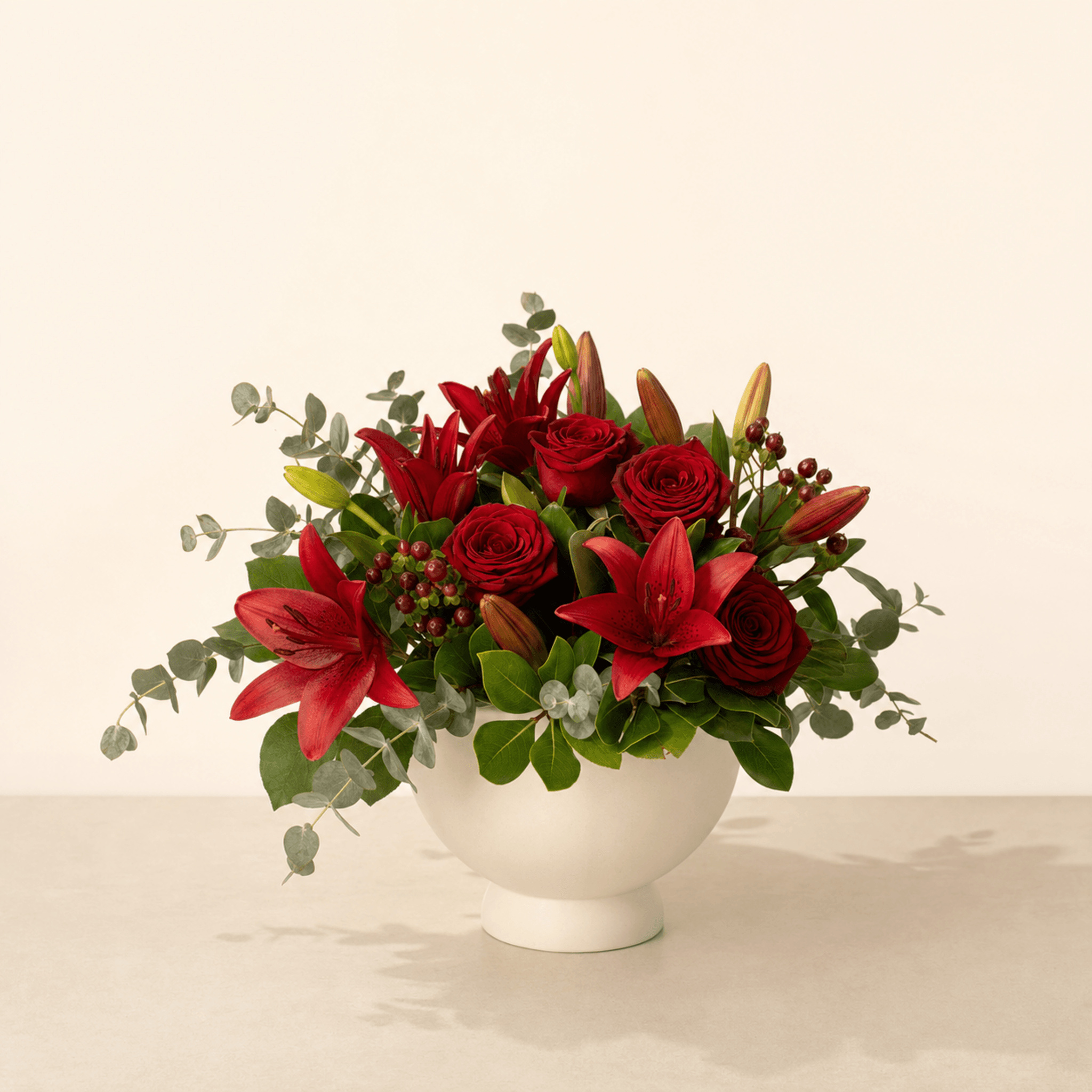Deep red roses and crimson lilies with eucalyptus and hypericum berries in a white ceramic bowl