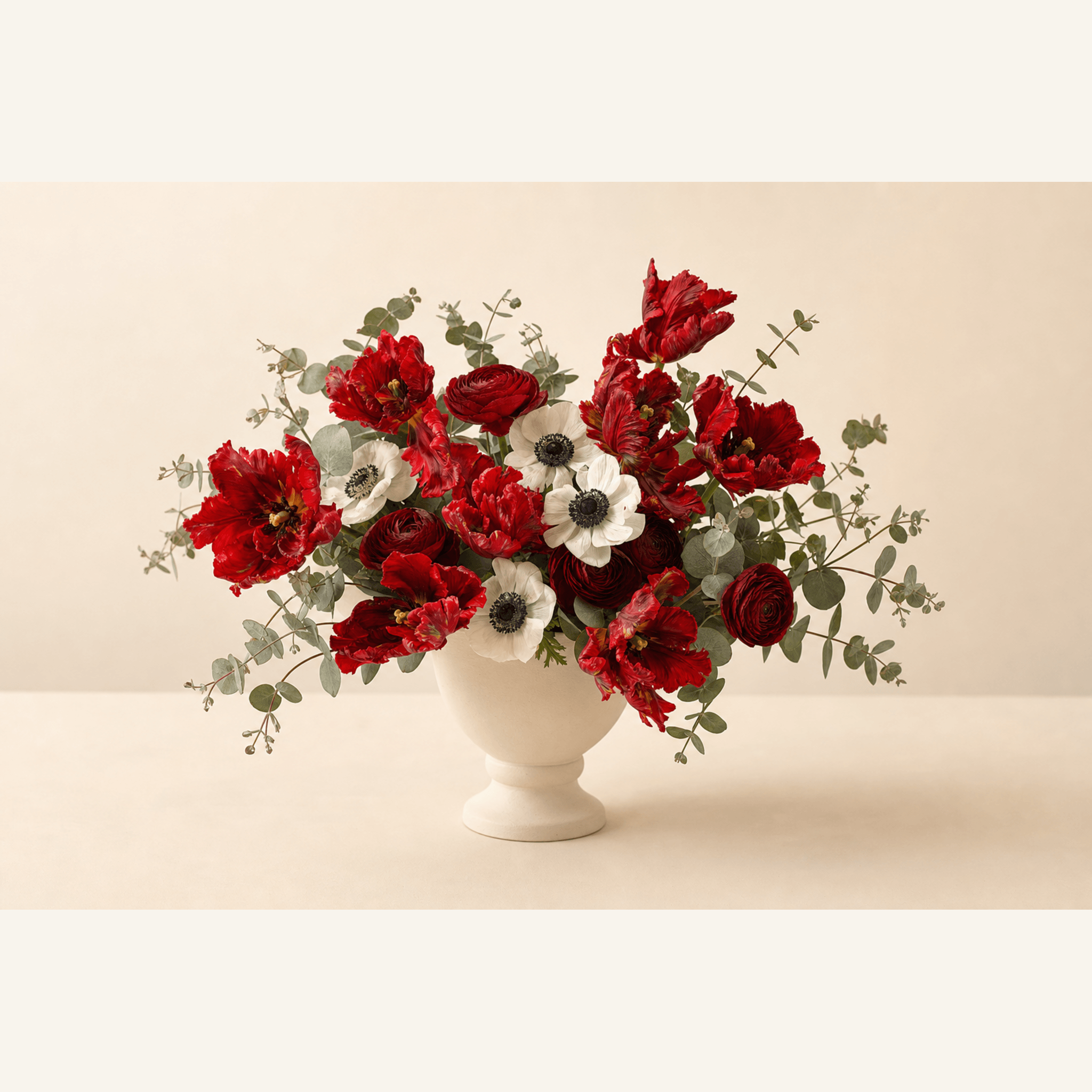 Red parrot tulips, red ranunculus and white anemones arranged in a white pedestal bowl with eucalyptus