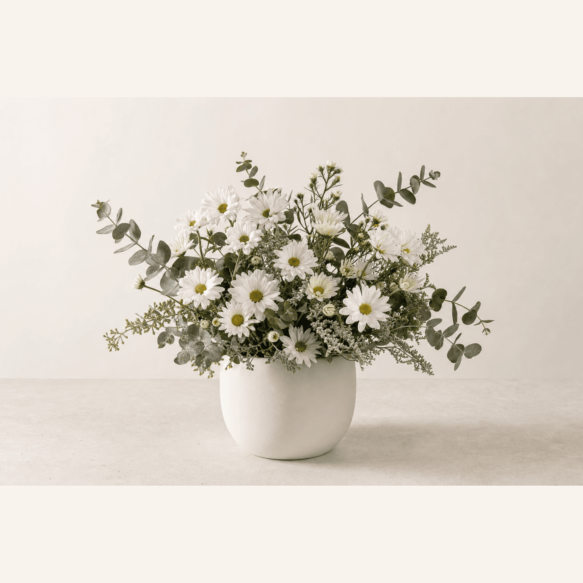 White daisies and eucalyptus sprigs arranged in a round white ceramic vase