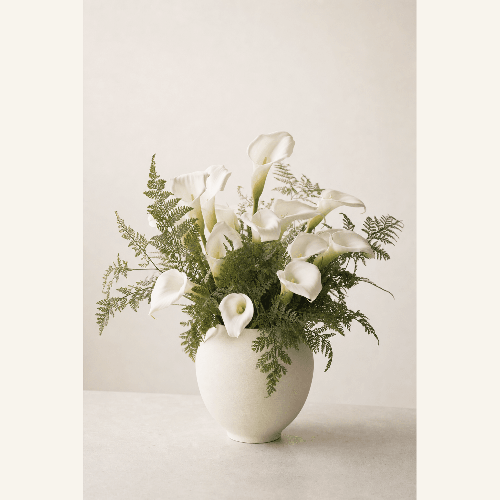 White calla lilies and soft green ferns arranged in a round cream ceramic vase