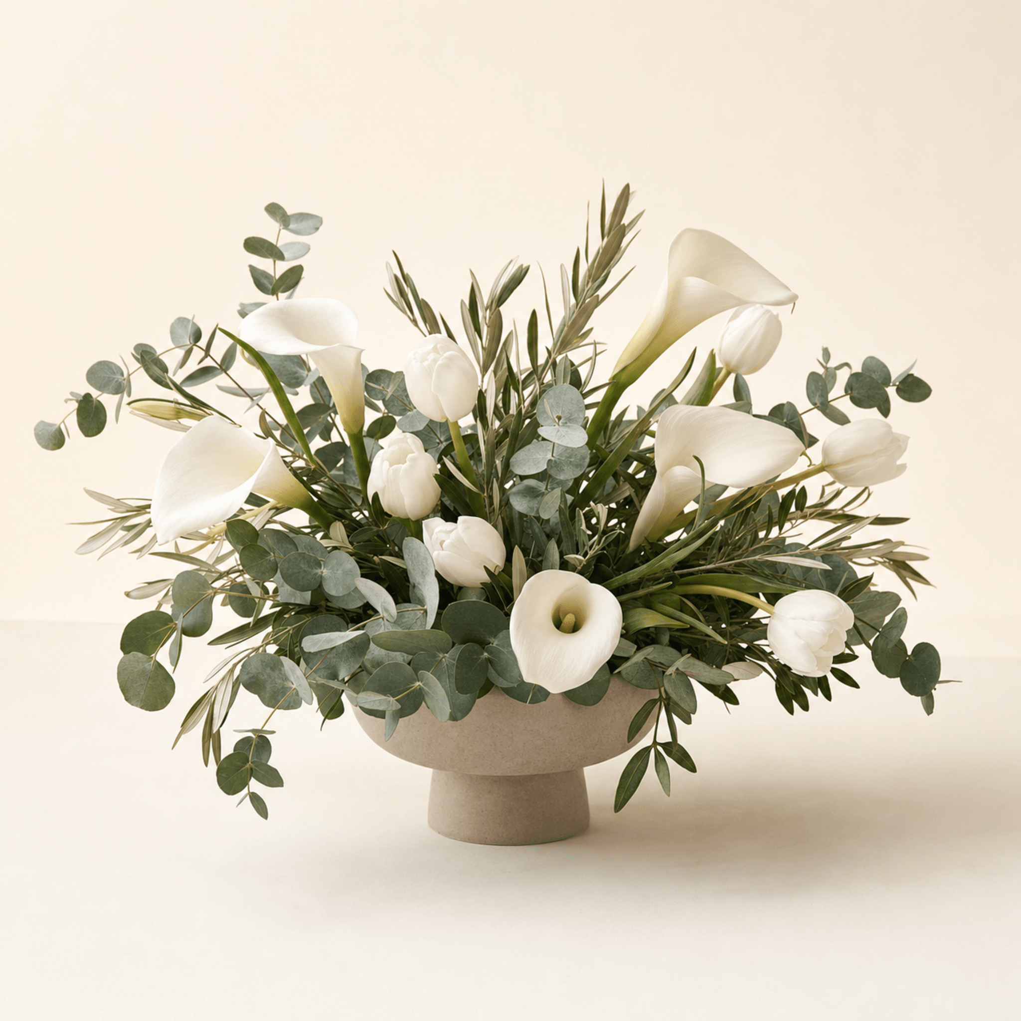 White calla lilies and white tulips with eucalyptus and olive foliage in a stone pedestal bowl