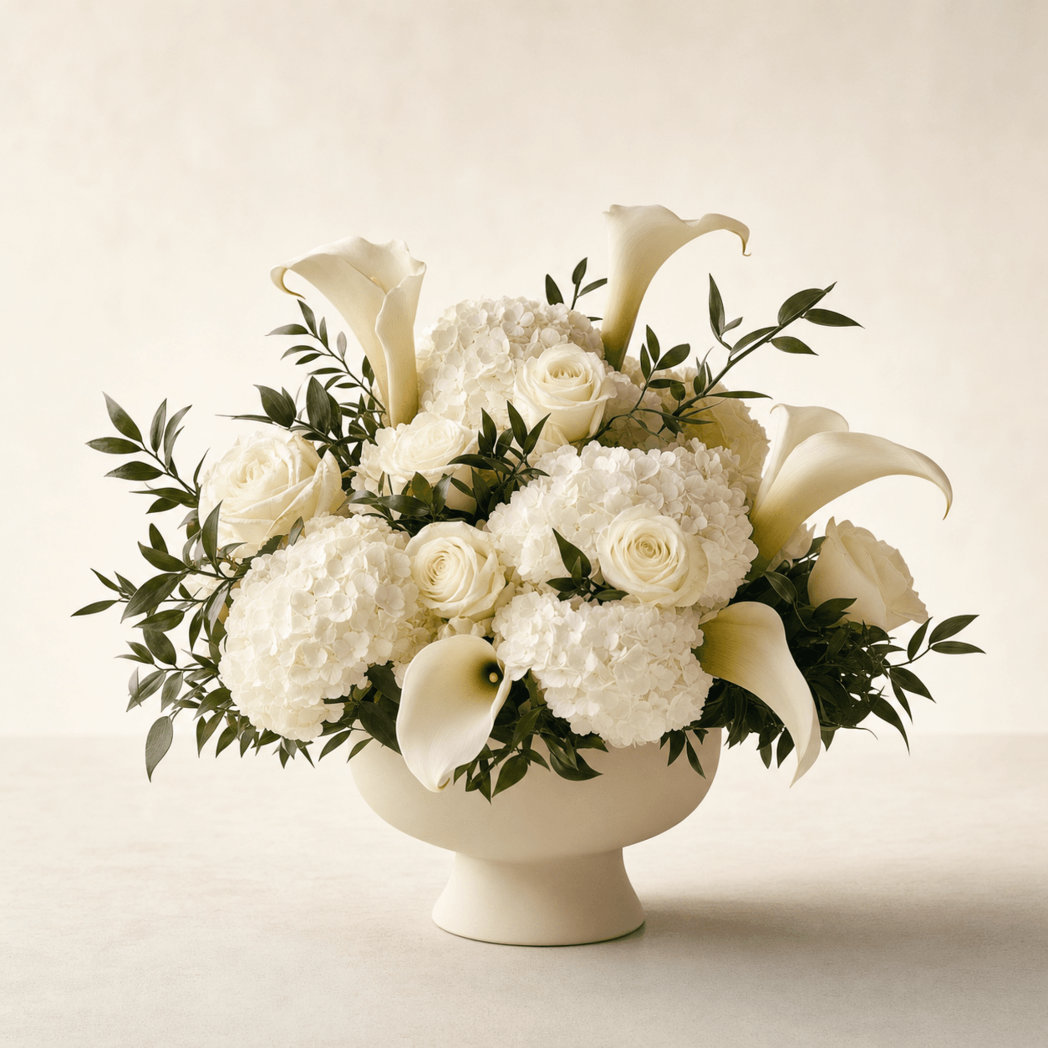 White calla lilies, cream roses, and white hydrangeas arranged in a cream ceramic pedestal bowl