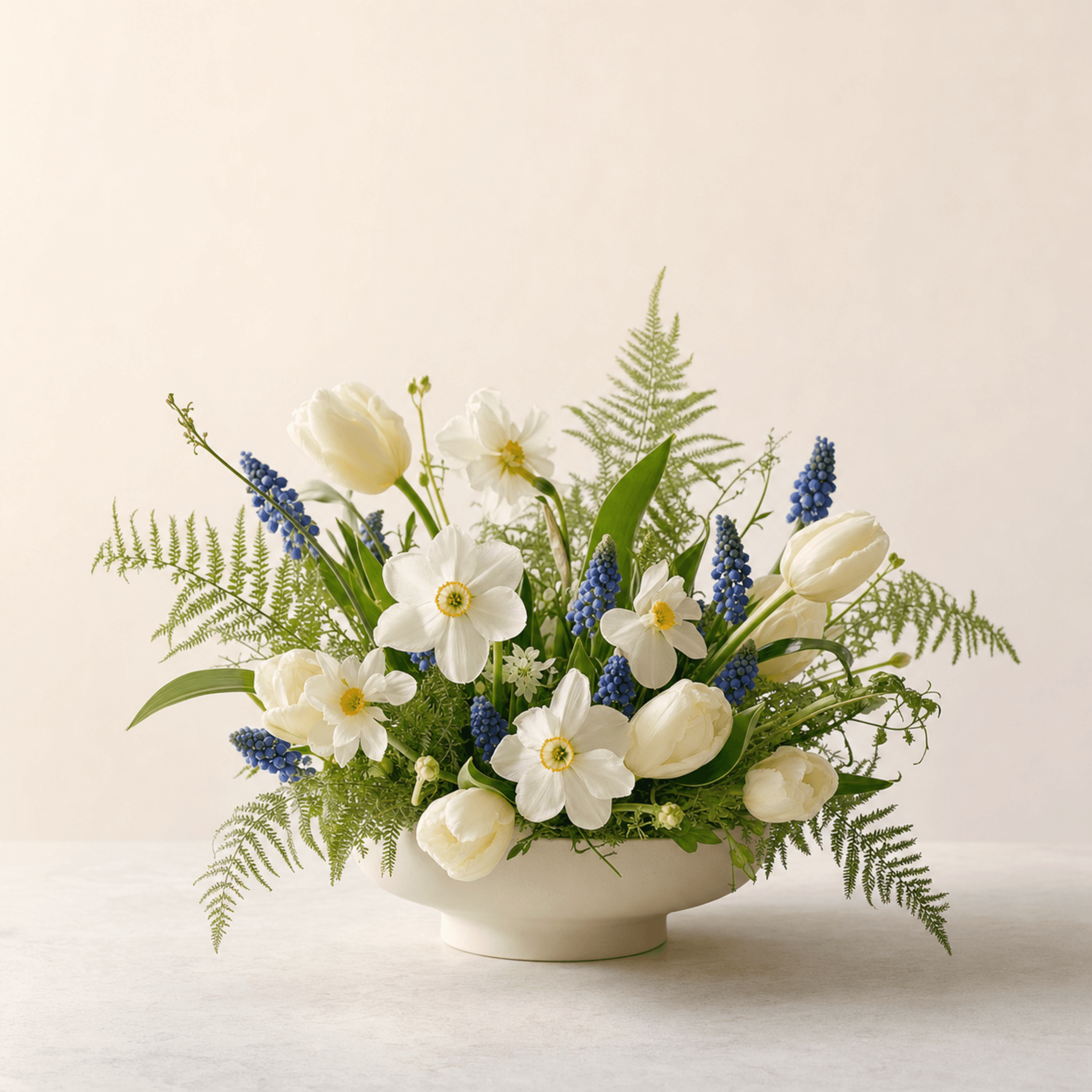 White tulips, narcissi, and blue muscari with fern fronds in a low cream ceramic bowl