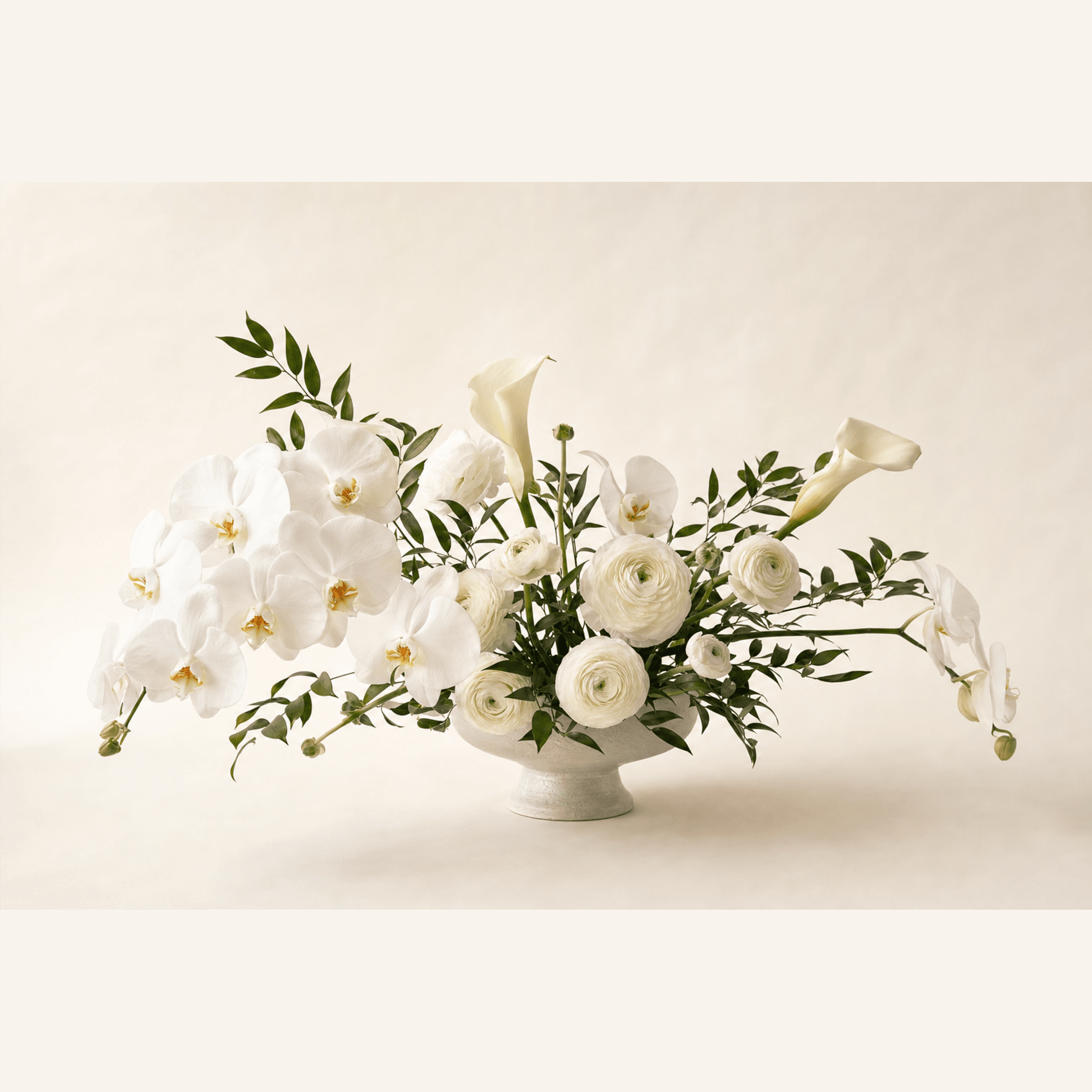 White phalaenopsis orchids, ranunculus and calla lilies with trailing green foliage in a white ceramic footed bowl