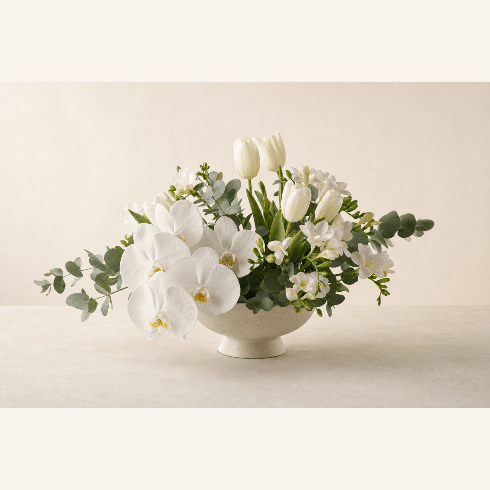 White phalaenopsis orchids, tulips and freesias arranged in a cream ceramic footed bowl with eucalyptus