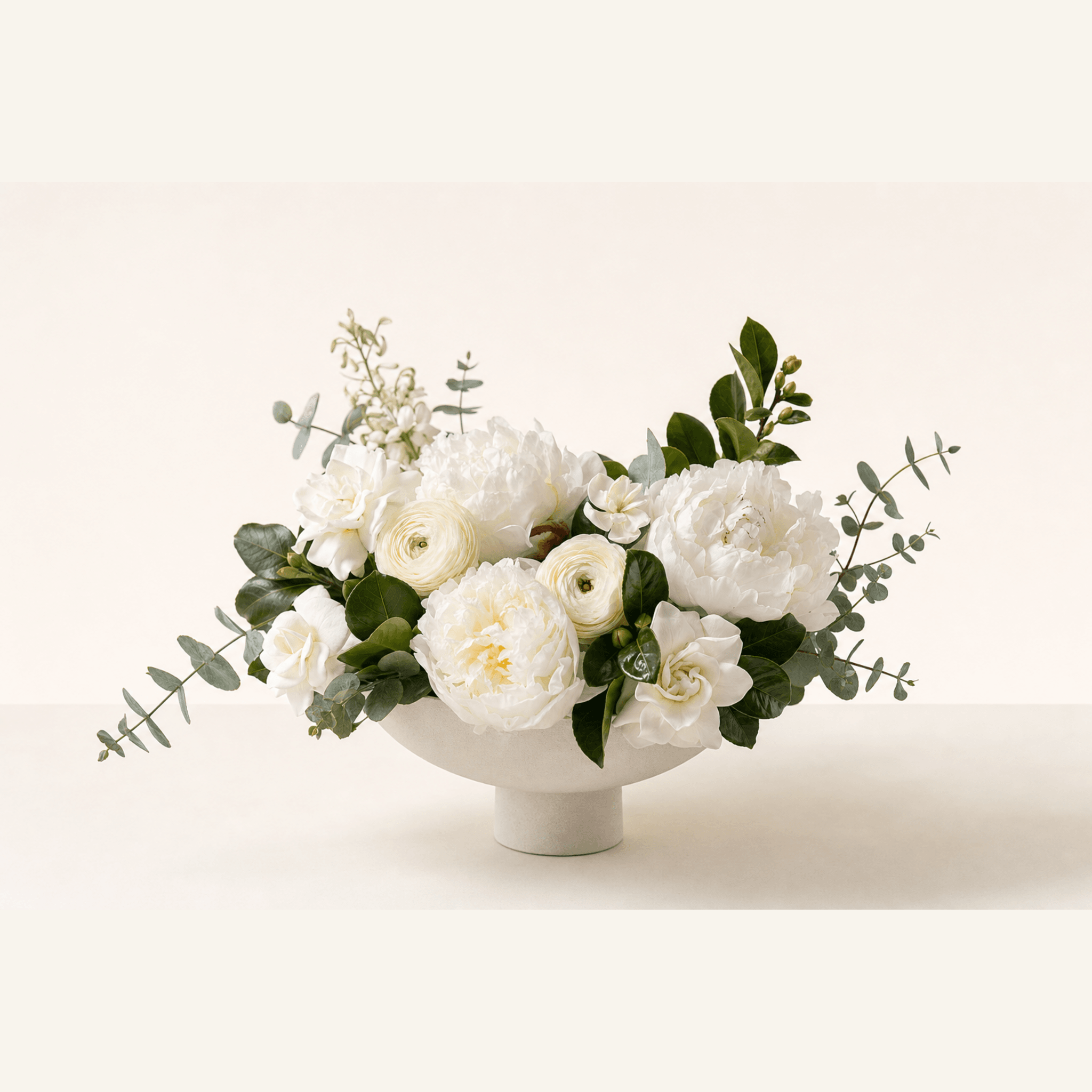 White peonies, ranunculus, gardenias and eucalyptus arranged in a white ceramic pedestal bowl