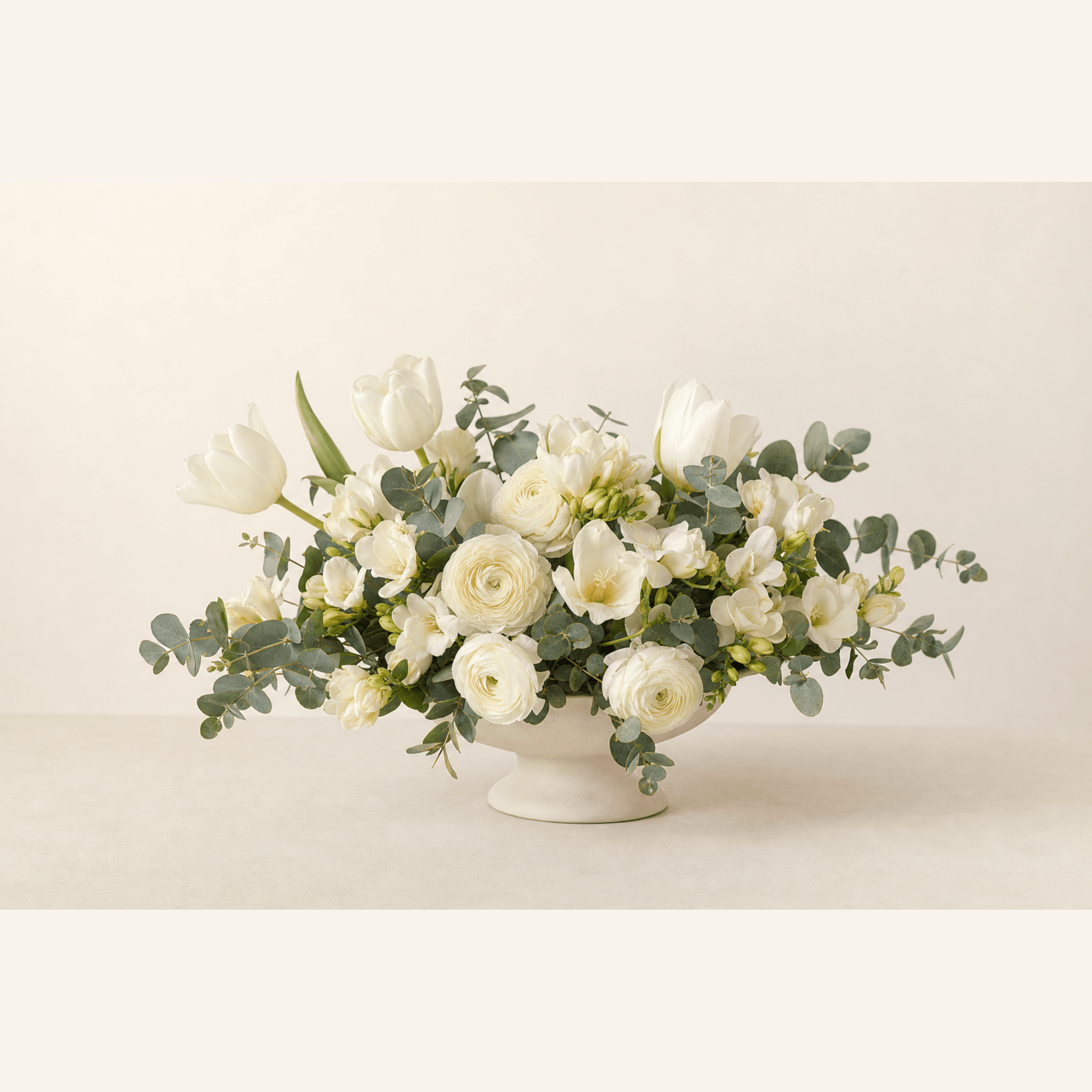 White tulips, ranunculus and freesia with eucalyptus in a white ceramic pedestal bowl