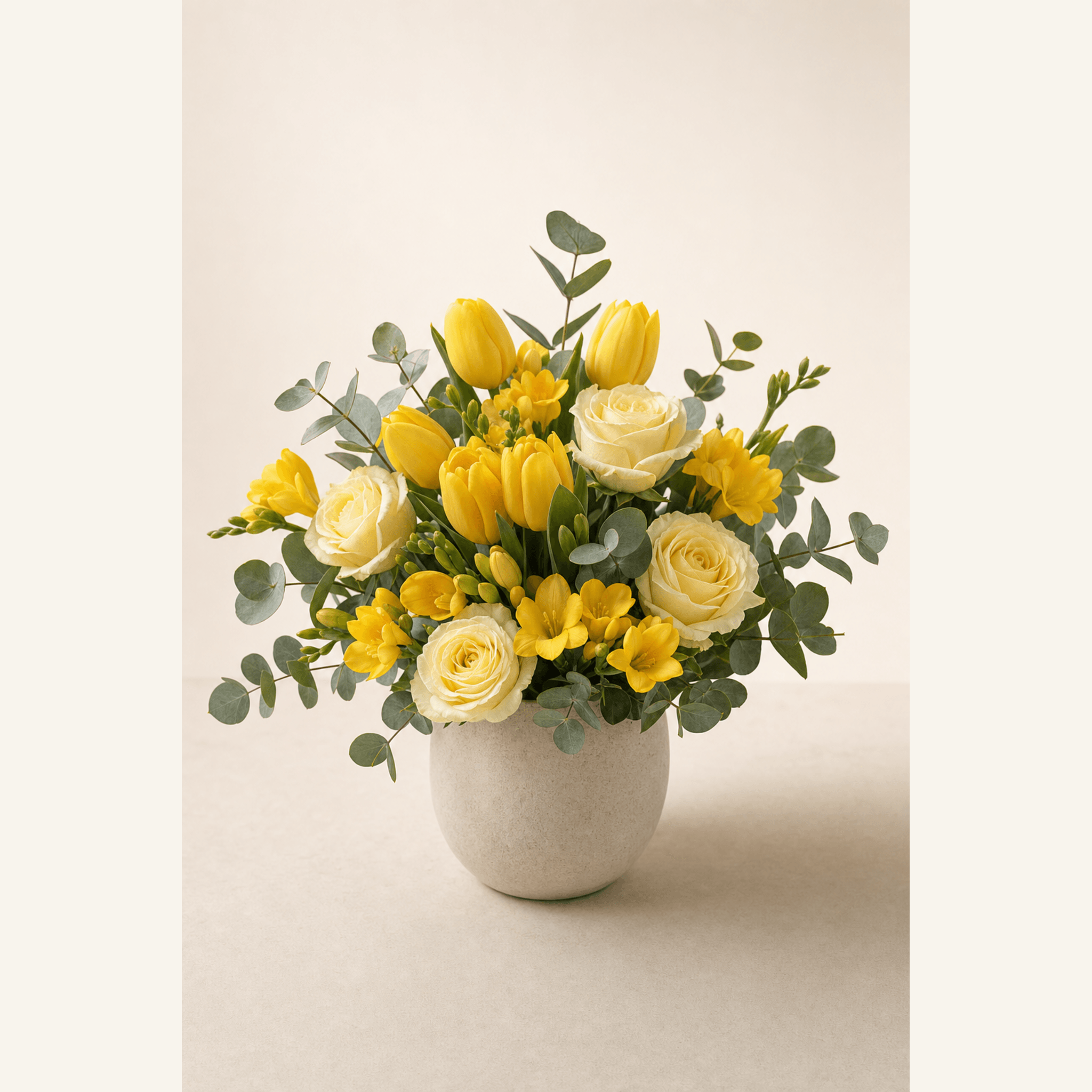 Yellow tulips, cream roses, yellow freesias and eucalyptus in a round stone vase